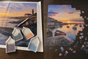 Een diamond painting met steentjes en bakjes naast een half gelegde fotopuzzel van een haven bij zonsondergang